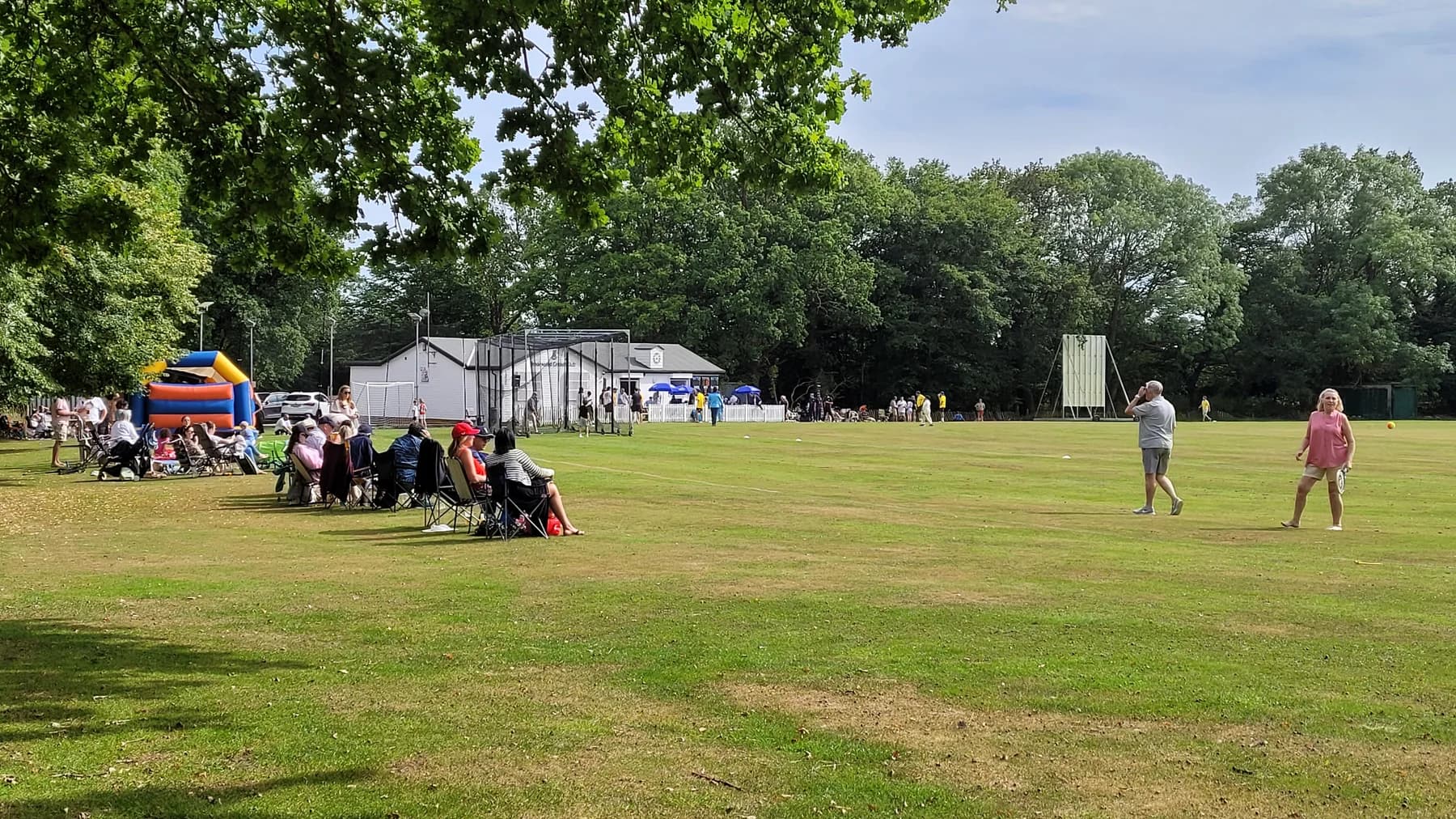 Spectators in deckchairs watching cricket beside the Brookweald clubhouse and practice nets on a summer afternoon